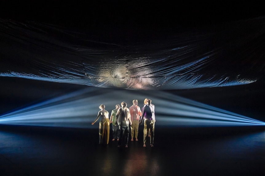 Five dancers are in the centre of a darkened stage spliced by beams of blue light, with a reflective sheet hanging above them