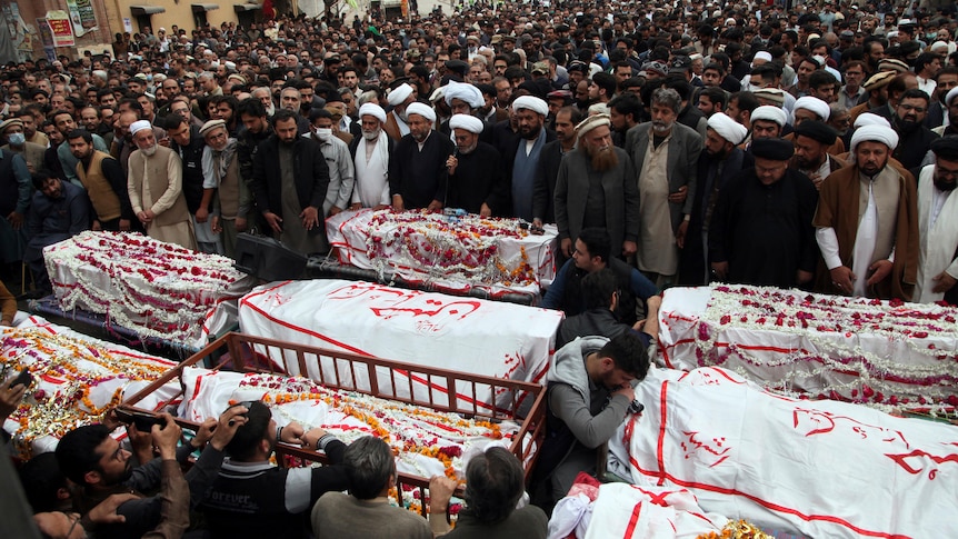 Hundreds of people stand beside coffins covered in white cloth and flower petals.