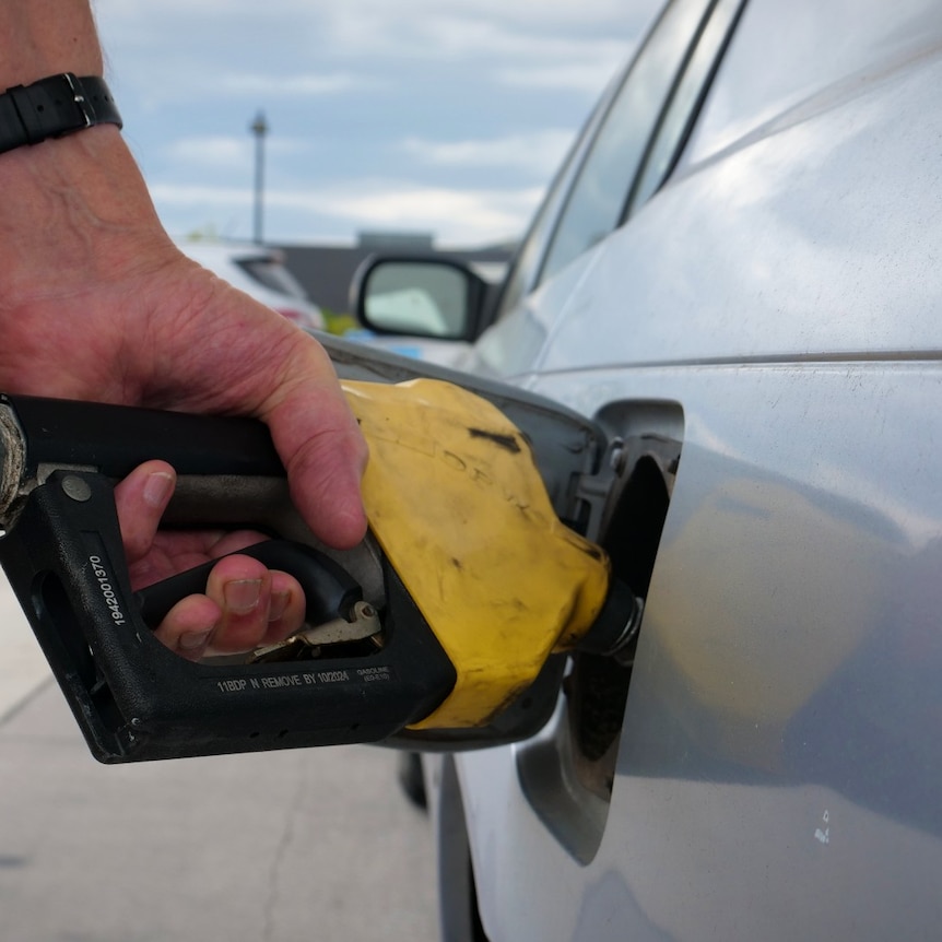 a hand with a watch holds a yellow petrol pump to a silver car