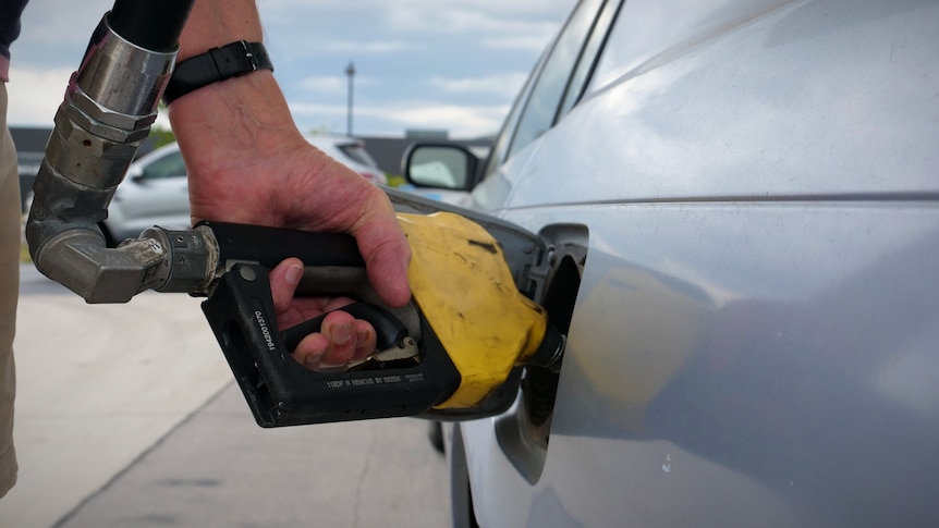 a hand with a watch holds a yellow petrol pump to a silver car