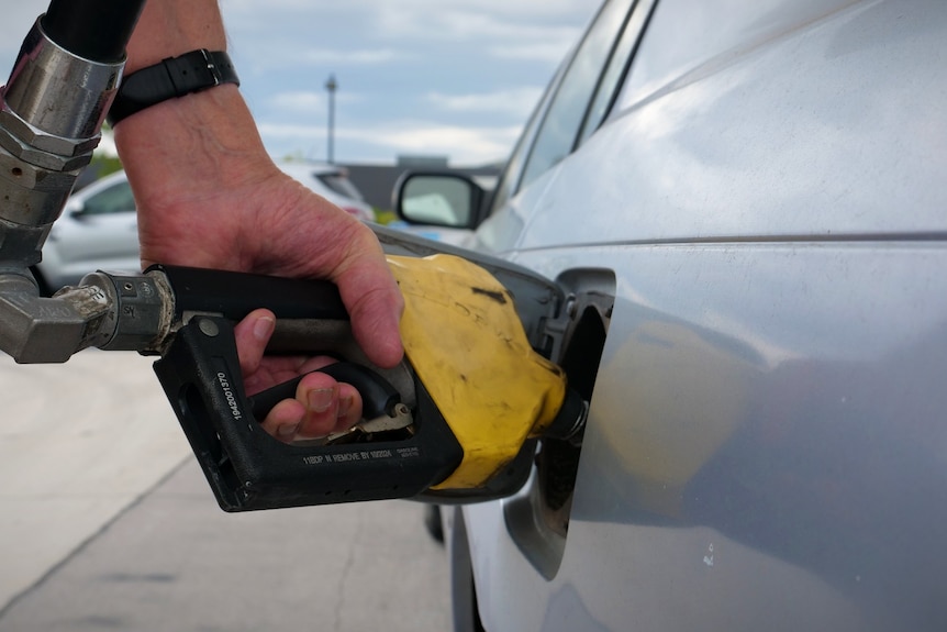a hand with a watch holds a yellow petrol pump to a silver car
