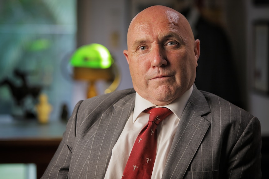 A man is seated indoors wearing a grey suit, white shirt and red tie.