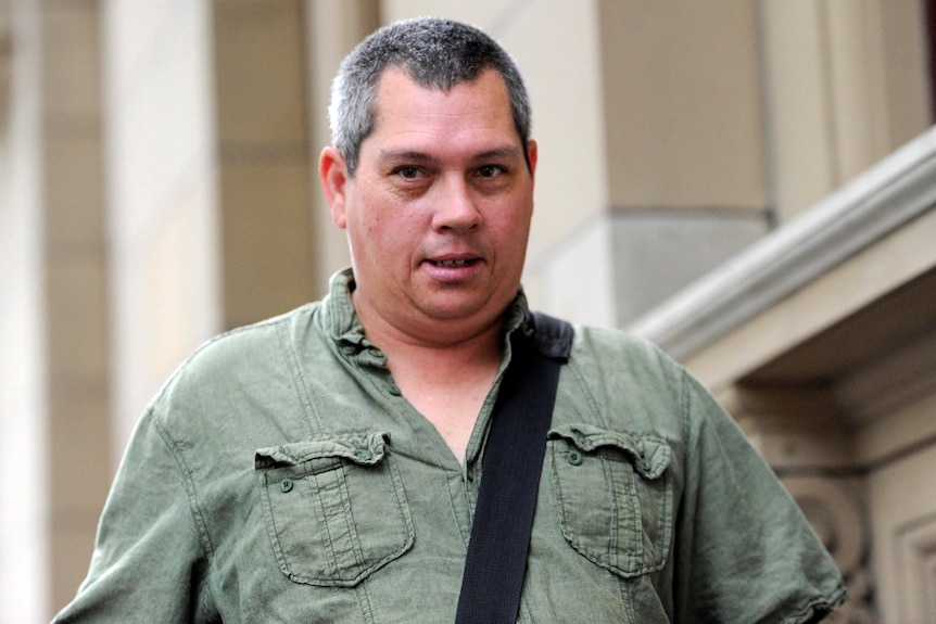 Brendan Sokaluk, a man wearing a khaki-green shirt and carrying a bag over one shoulder, looks past the camera.