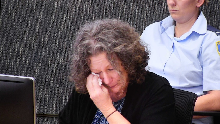 A woman wearing a black cardigan sitting down wipes her eyes.