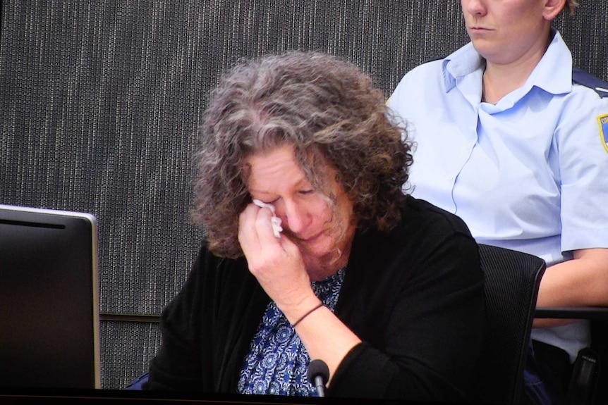 A woman wearing a black cardigan sitting down wipes her eyes.