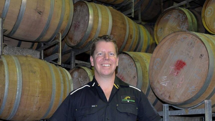 A man in a dark shirt smiles while standing in front of a bunch of wine barrels.