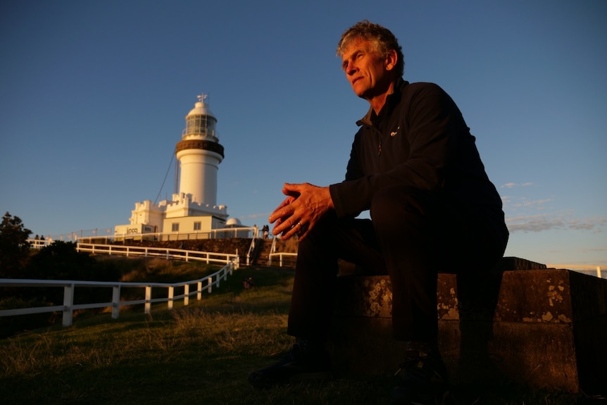 A man in a long black shirt sitting on a concrete block with a lighthouse in the background