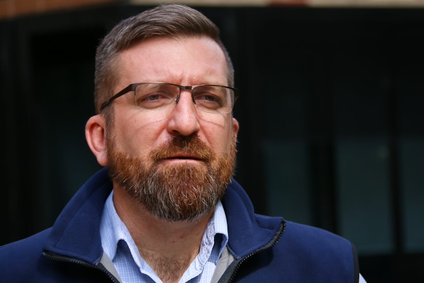 A man with glasses and a puffer jacket stands outside an office building.