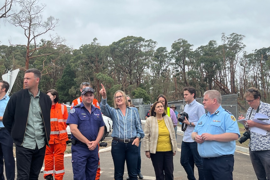 Jacinta Allan wears a blue shirt and jeans and points off camera while standing beside Lily D'Ambrosio and emergency workers.