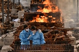 Two people dressed in blue scrubs and wearing masks lean on each other and cry in front of a fire.