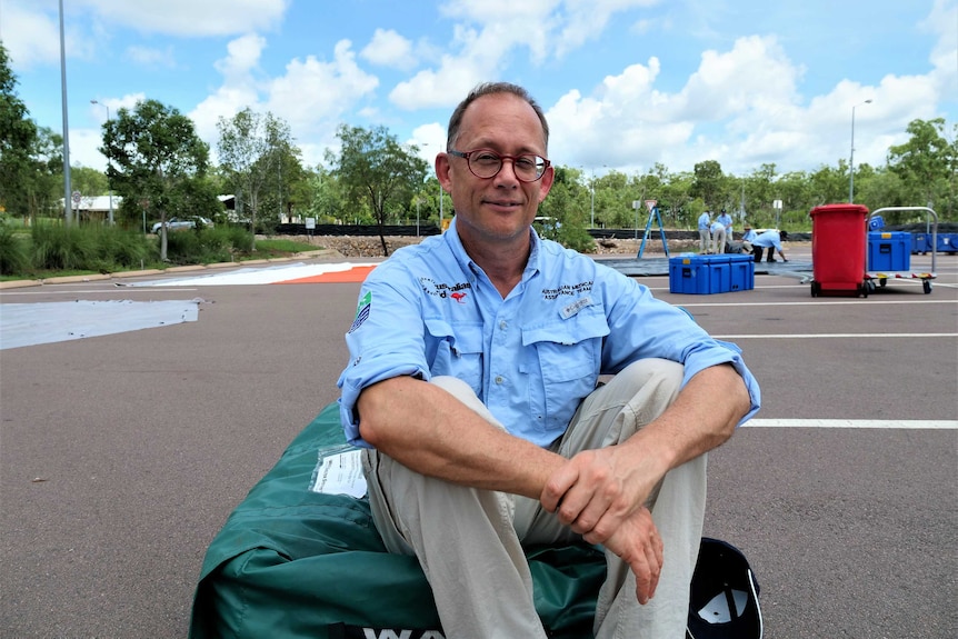 A man wearing the blue Ausmat shirt in a carpark sitting on a packed-up tent sunny day clouds.