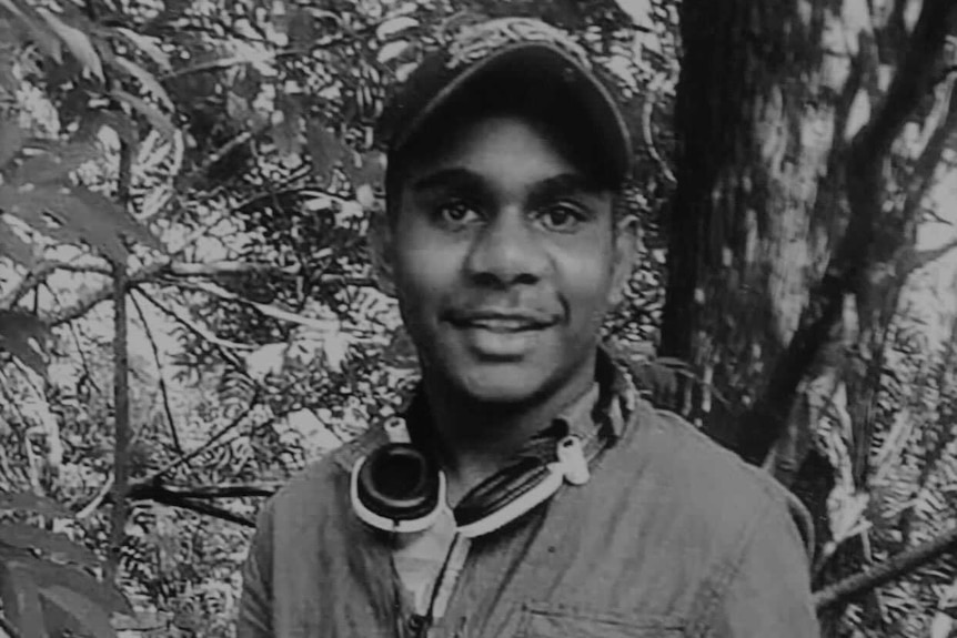 A black and white image of a young man smiling, wearing a baseball cap, headphones around his neck