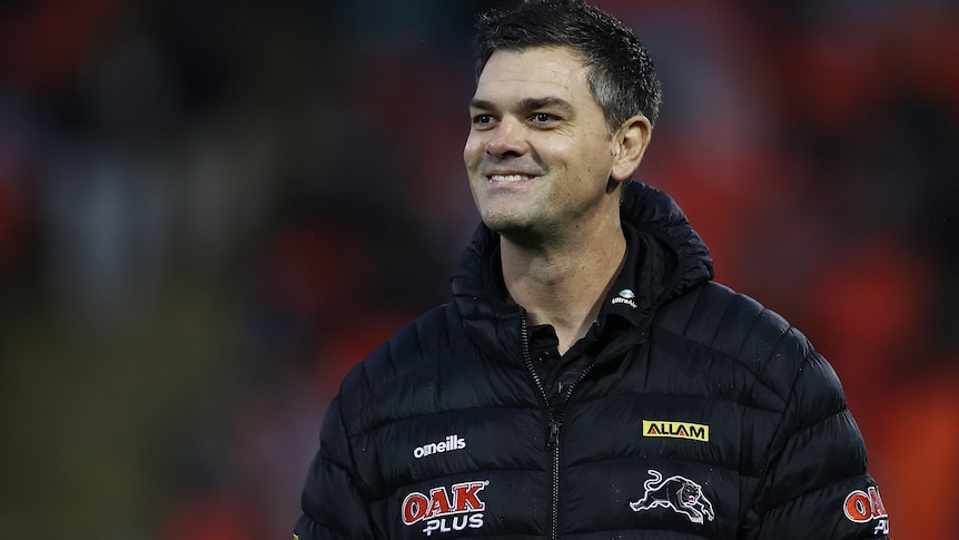An assistant coach wearing club gear with the Penrith Panthers logo smiles as he looks across a field before a game. 