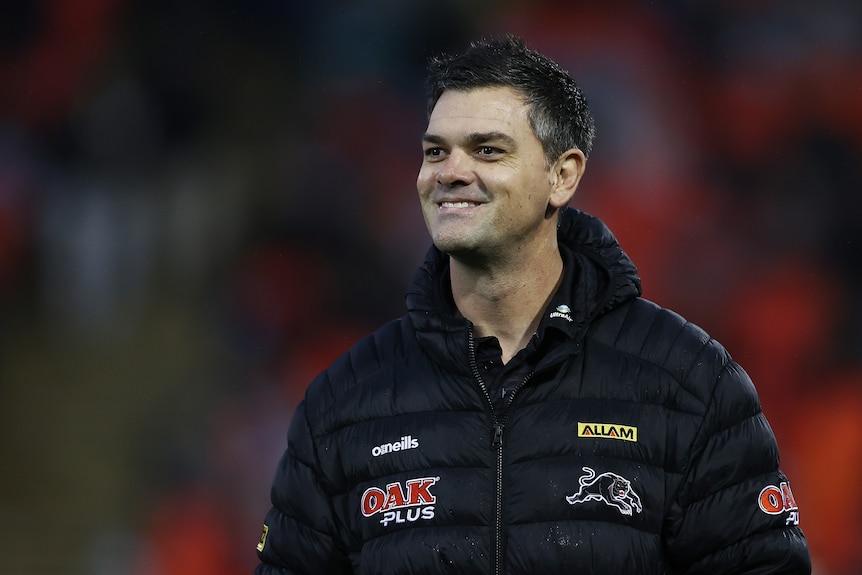 An assistant coach wearing club gear with the Penrith Panthers logo smiles as he looks across a field before a game. 