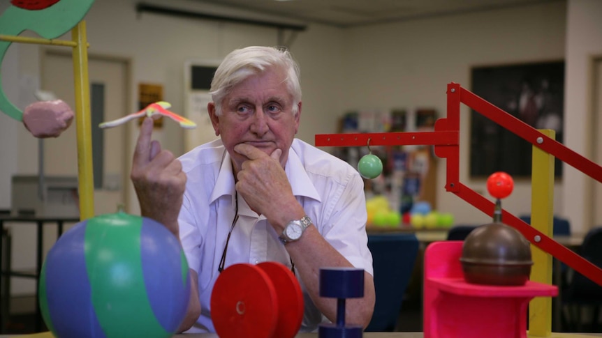 Scientist balances a toy bird on his finger and sits at a desk with various colourful props.