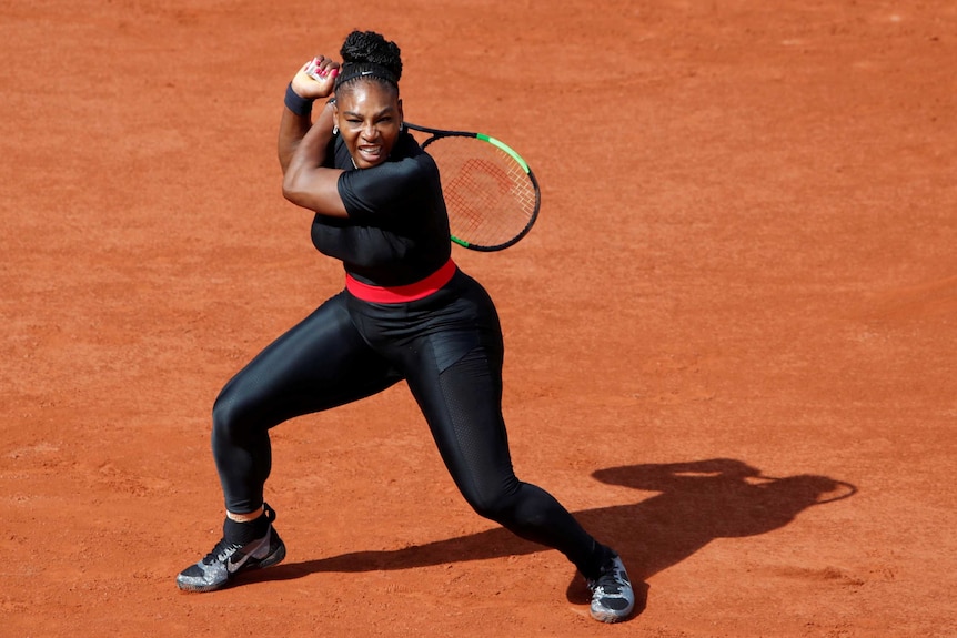 Serena Williams swings her racket over her shoulder, behind her back, half crouched, wearing a skin-tight black suit.