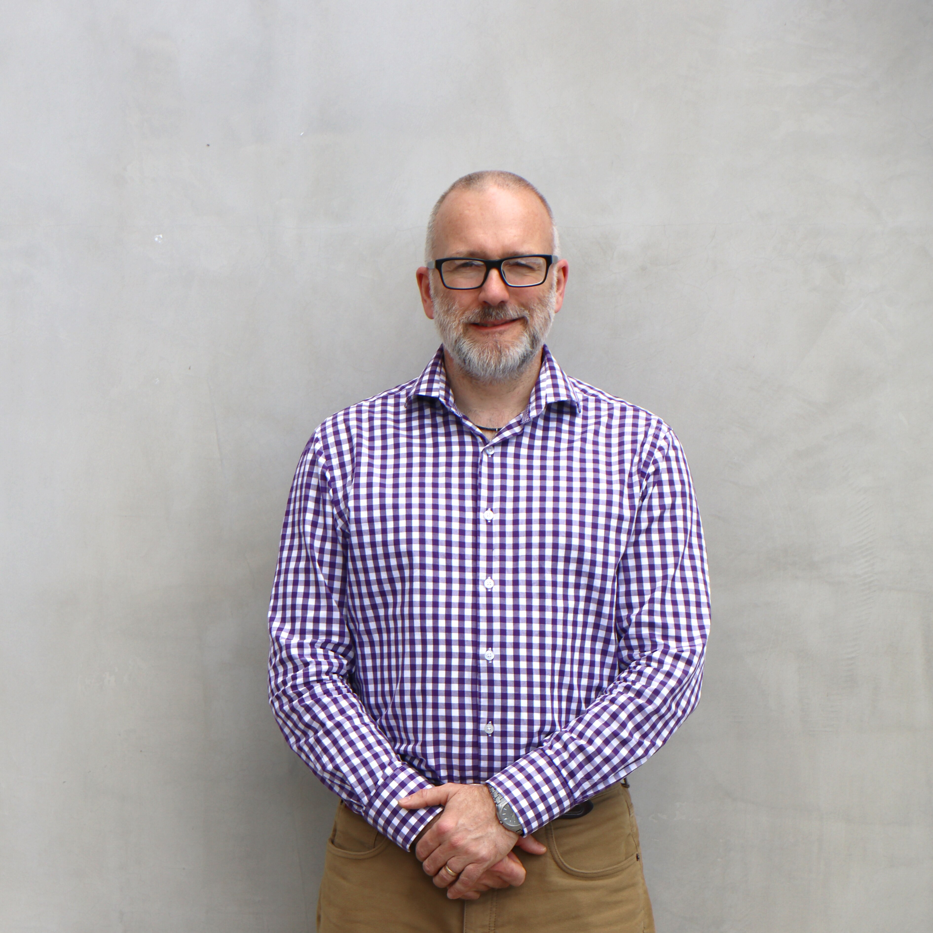 A man with grey hair and glasses wearing a purple checked shirt.