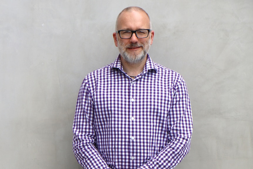 A man with grey hair and glasses wearing a purple checked shirt.