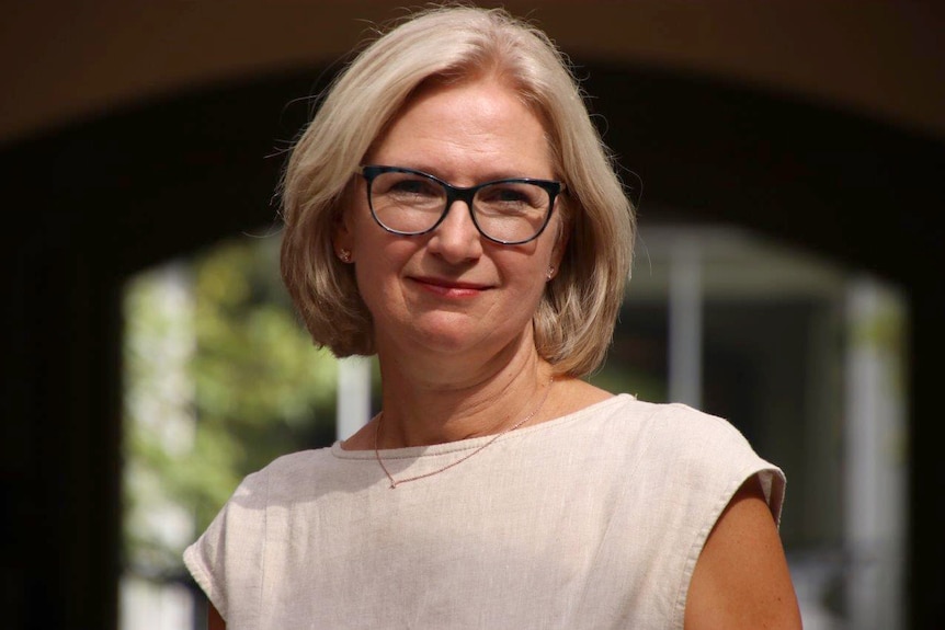 A head and shoulders shot of a middle aged woman with blond hair and black-framed spectacles posing for a photo outdoors.