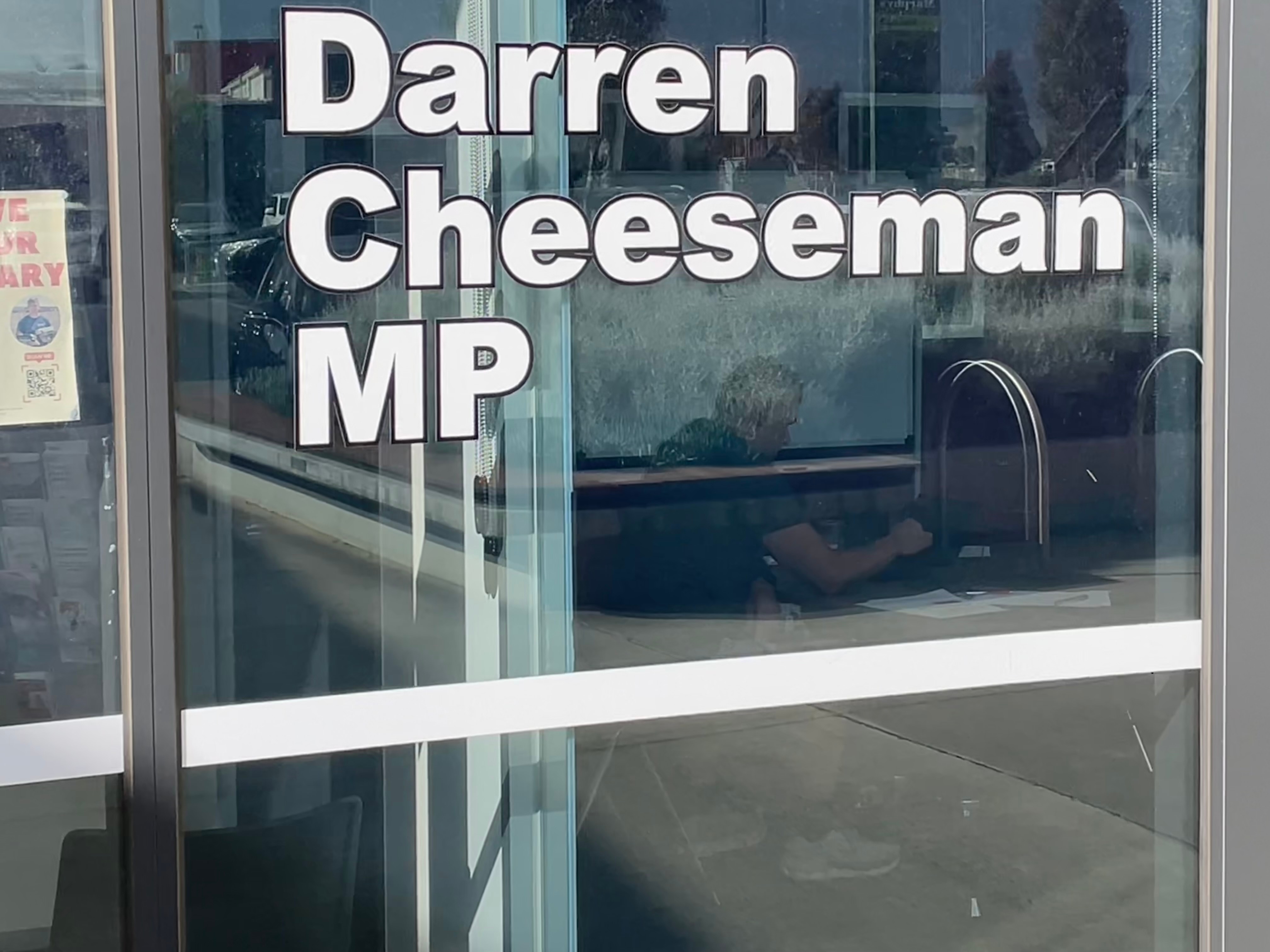 A man is visible through a window sitting at a desk, with a large sign printed on the window which says Darren Cheeseman MP
