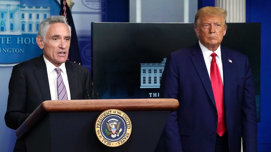 Atlas stands at the White House lectern speaking, as Trump stands off to the side