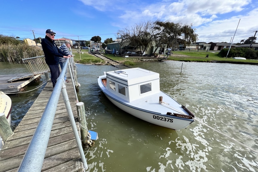 Two men stand on a narrow jetty over the river as they look down at a restored white wooden boat