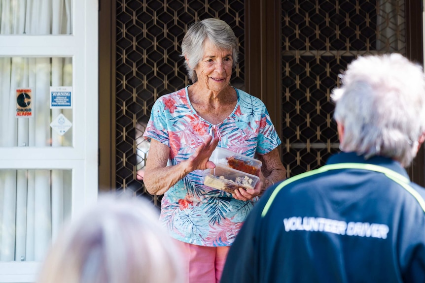 Older woman stands at front door holding meals in plastic boxes