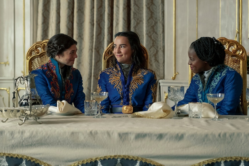 Three women wearing ornate blue jackets sit at a fancy dining table.