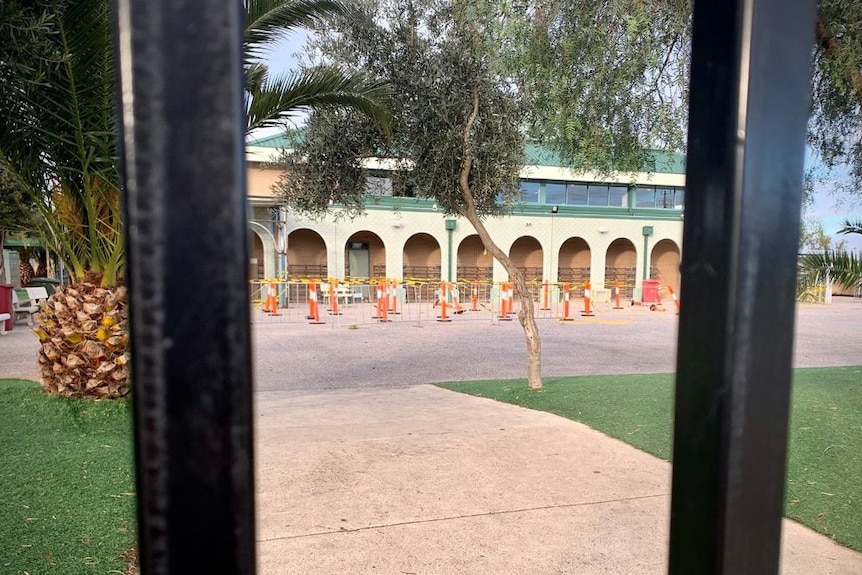 A photo taken through the school gates shows a large school building blocked off by bollards and yellow and black tape.