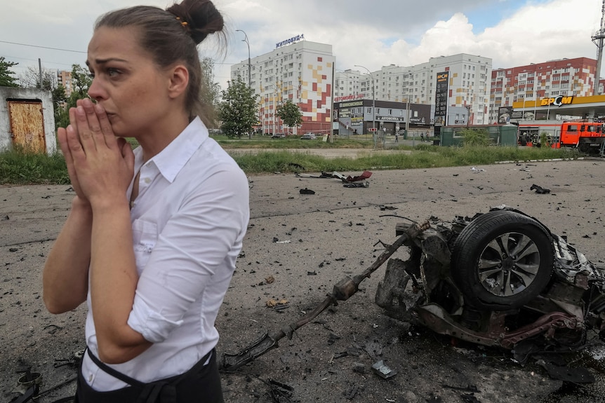 A woman covers her mouth as she stands behind the remains of a bombed car on a suburb street 