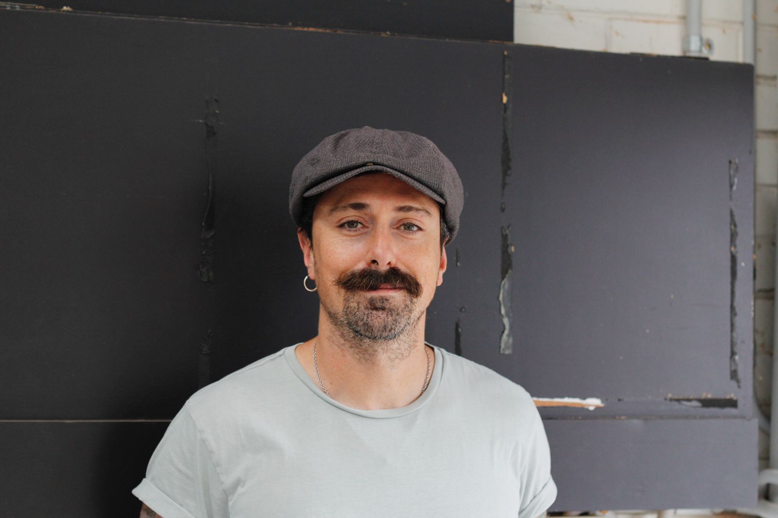 검은색 벽에 회색 평모자와 밝은 회색 티셔츠를 입은 남자의 사진이 있습니다.