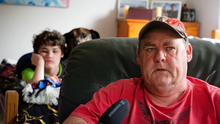 Man looking off in the distance while younger son and dog sit in background on a bed