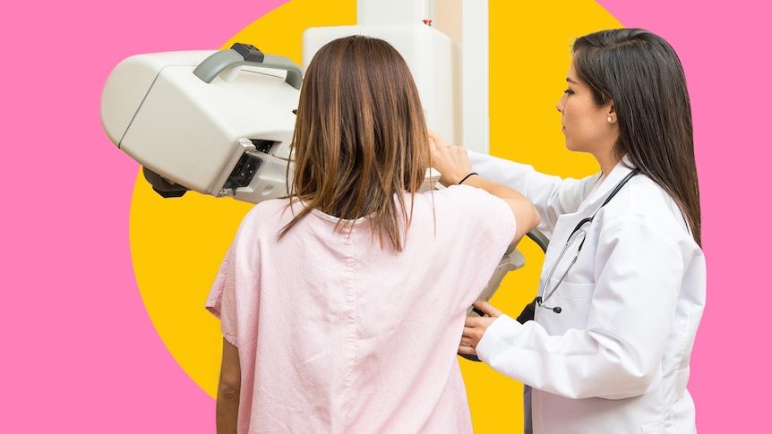 A woman, seen from behind as she stands in front of a mammogram scan machine with a doctor/radiographer lowering it toward her.