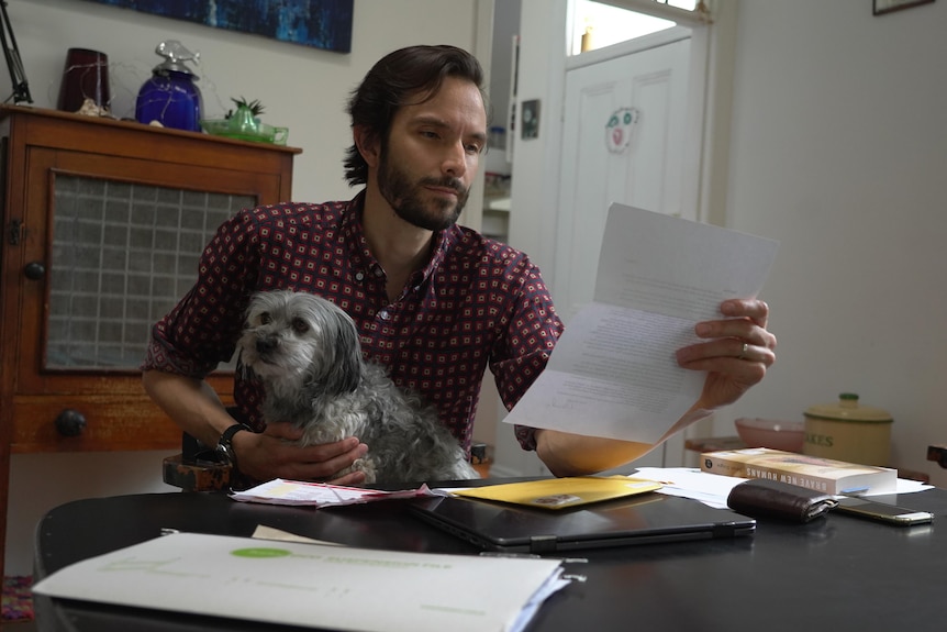 A young man sits at a desk holding letters in one dog and his small dog on his lap