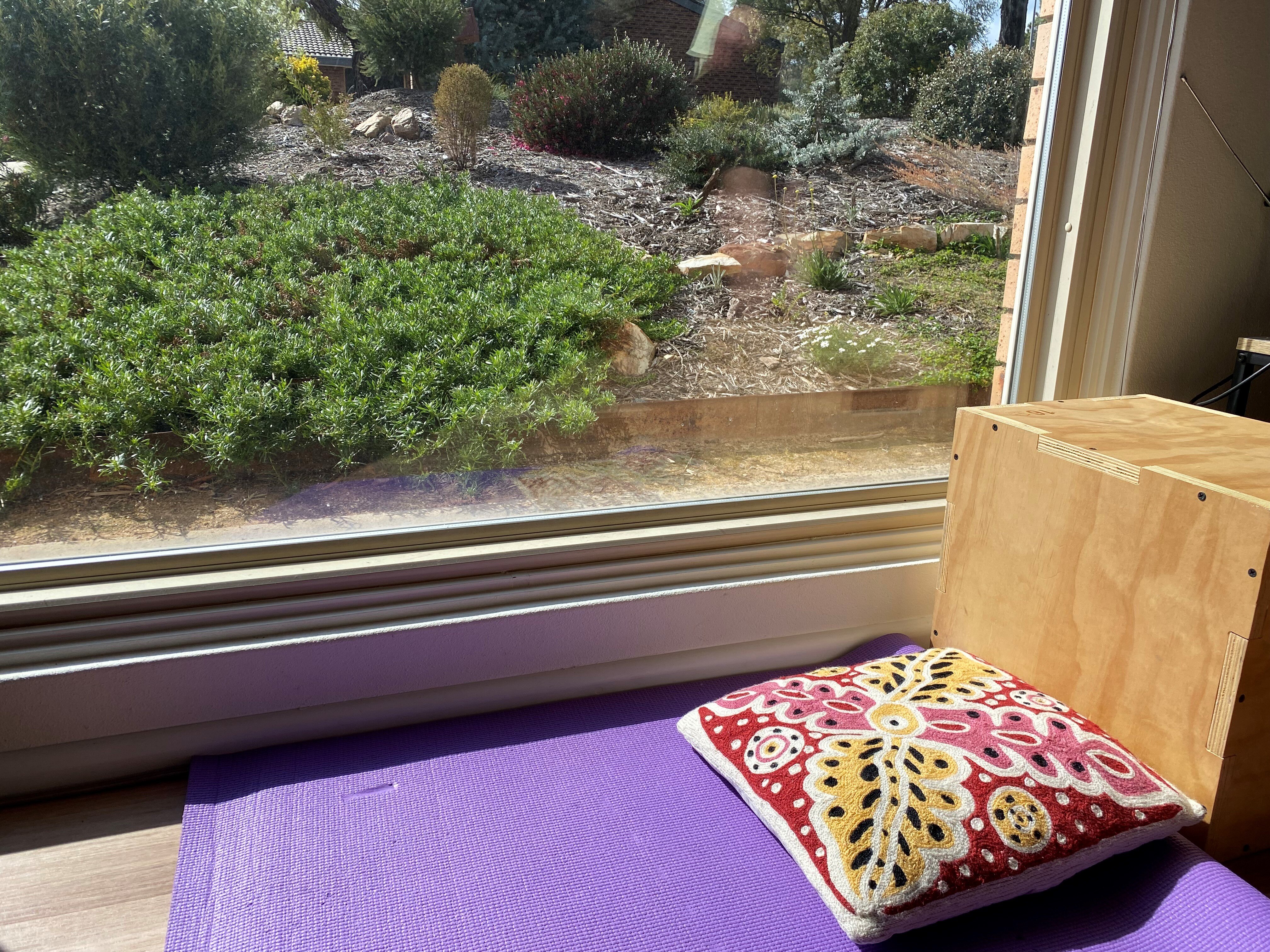 Коробка стоит на коврике для йоги с подушкой возле окна с видом на сад. Солнце входит в окно