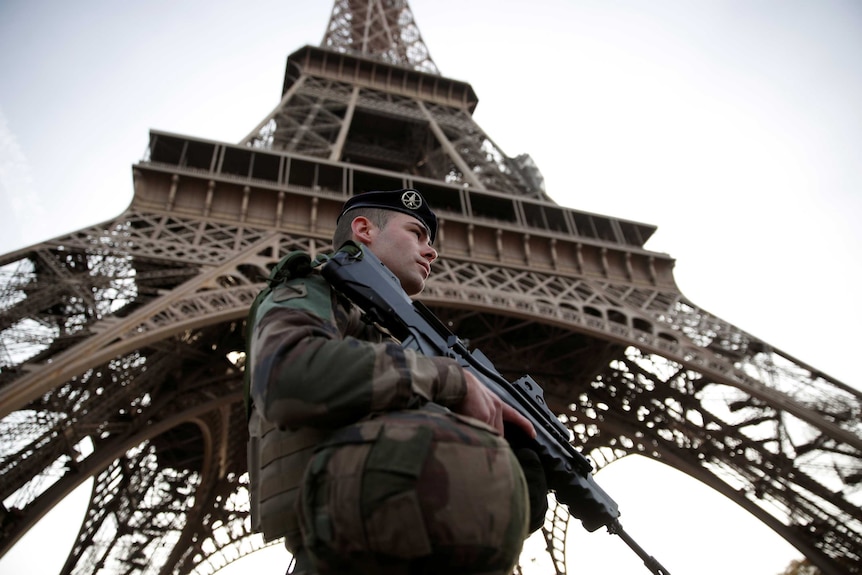 A French soldier, holding a high-powered gun, stands guard under the Eiffel Tower.