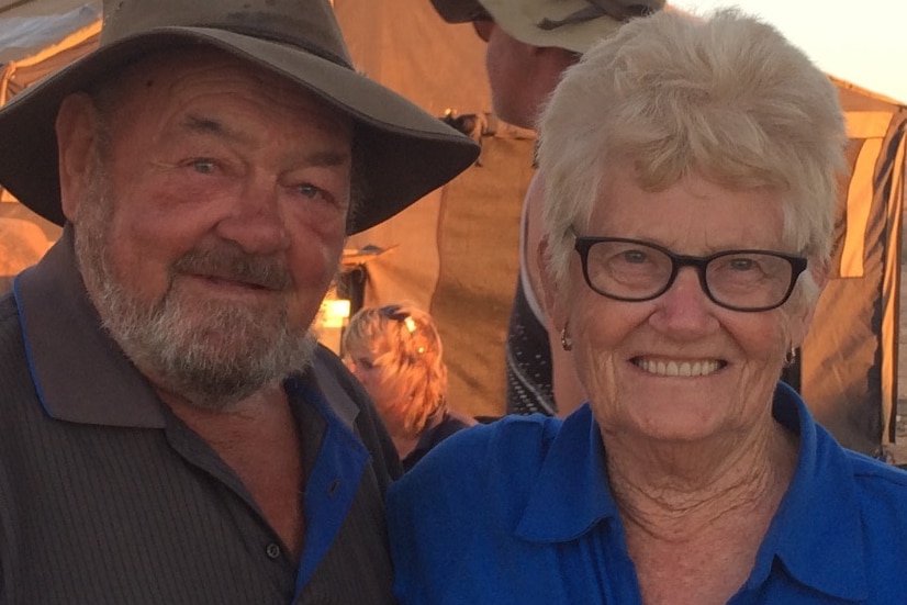 An elderly man in a brown-brimmed hat in front of a tent at sunset stands smiling next to an elderly woman in glasses.