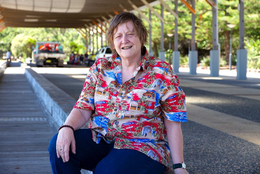 A smiling woman wearing a floral shirt, sitting in the shade of a large open-walled shed structure.