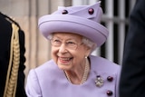 A close up of the Queen smiling in a purple coat and matching hat.