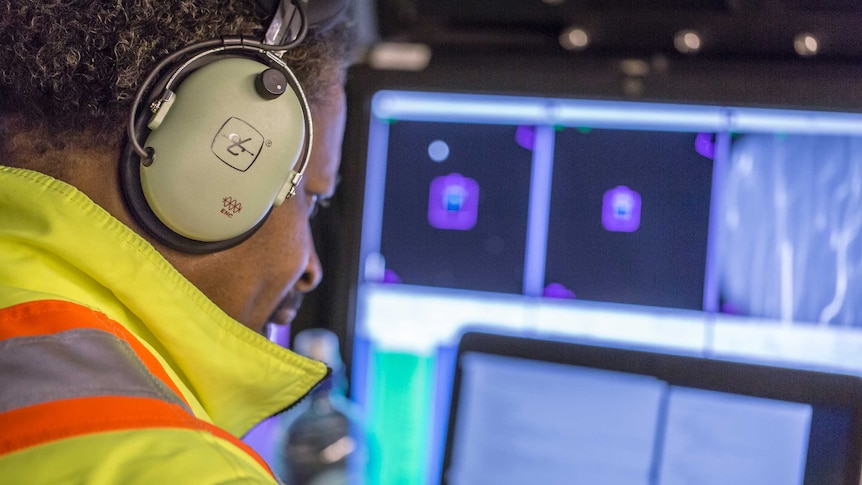 A man wearing headphones and a fluoro jacket looks at computer screens.