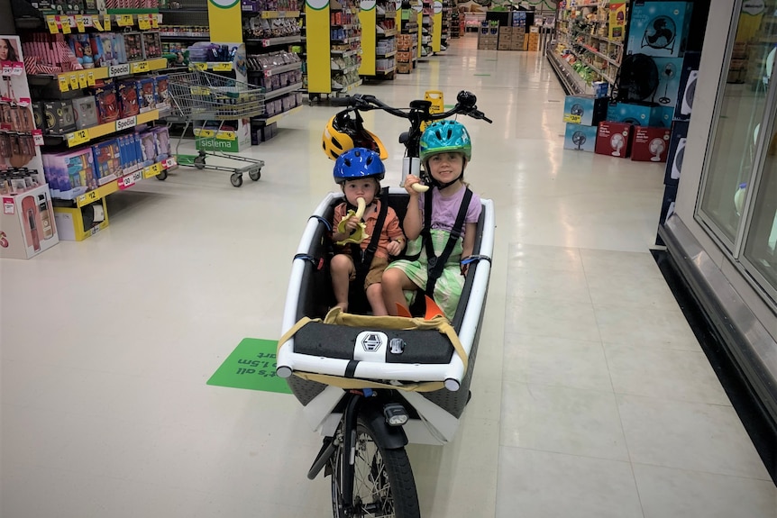 Two kids on a cargo box bike eating bananas in a supermarket aisle.