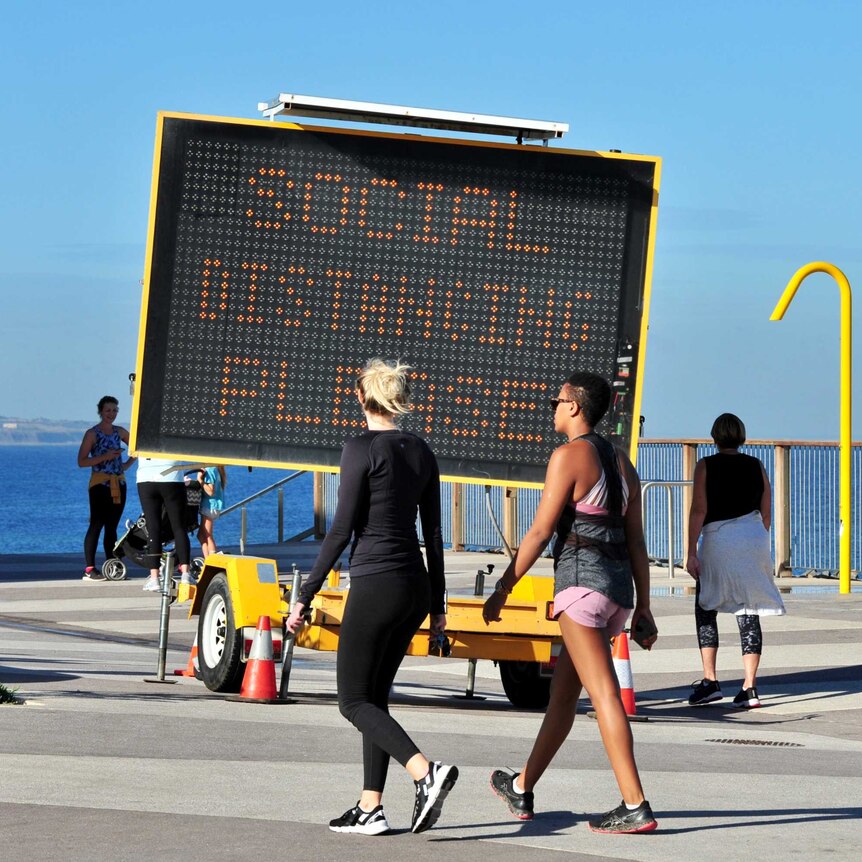 Two women walk in front of a social distancing LED sign on a boardwalk