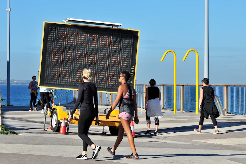 Two women walk in front of a socially distancing LED sign on a boardwalk
