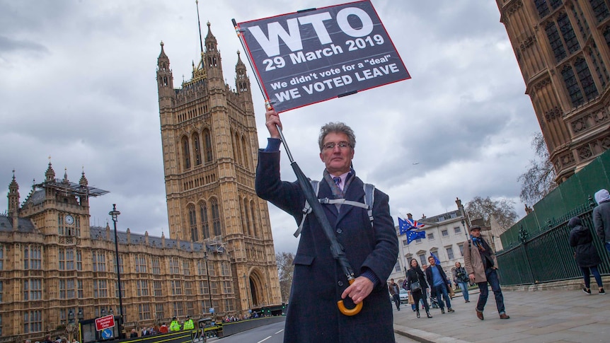 A man holding a protest banner attached to an umbrella stands outside Westminster.