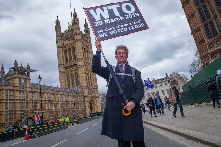 A man holding a protest banner attached to an umbrella stands outside Westminster.