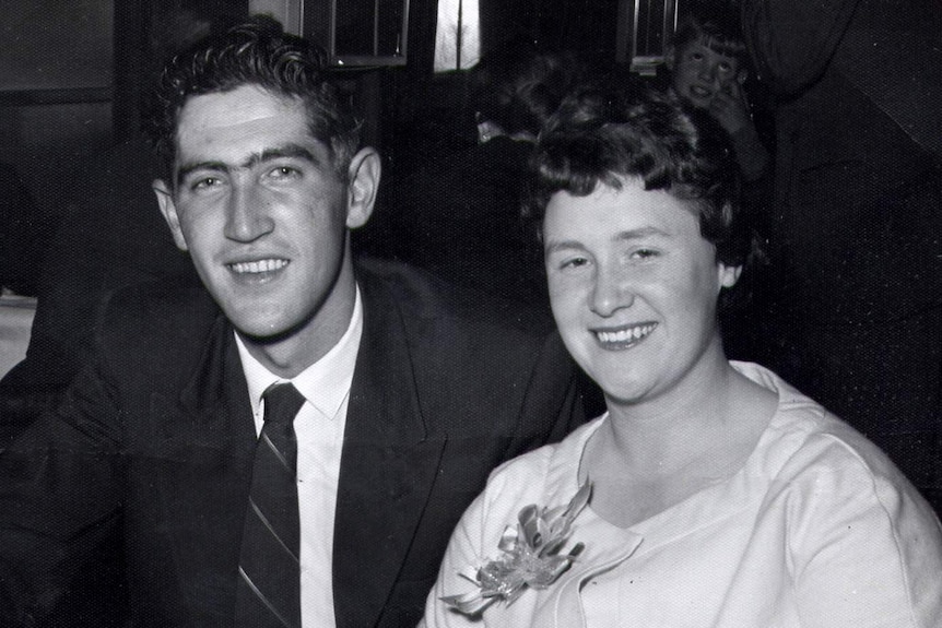 A young husband and wife smile in a black-and-white photo. The man wears a suit and tie, the woman wears a pale suit with brooch