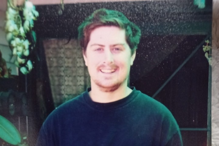 A man with brown hair and a goatee wears a black tshirt and smiles. 