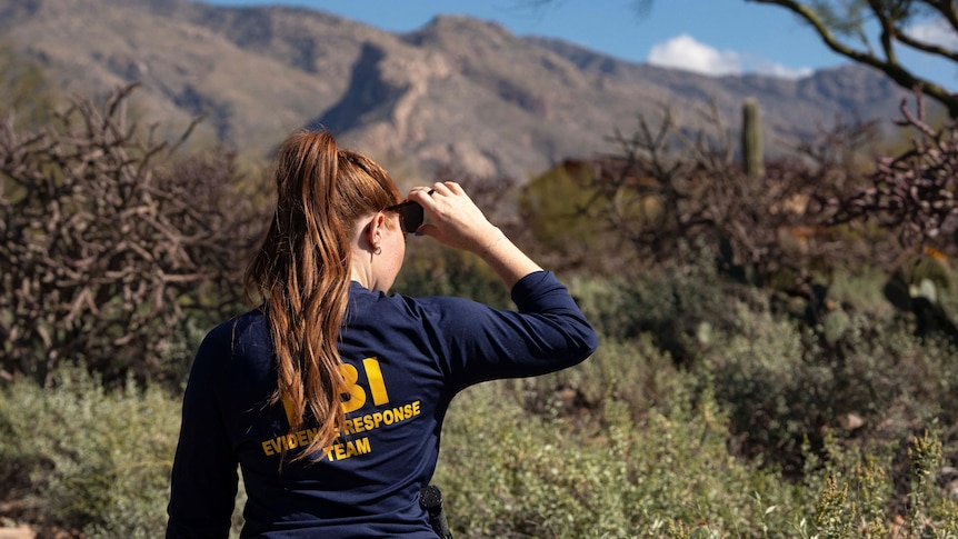 A woman with long, red hair wears an FBI shirt as she scans arid wilderness.