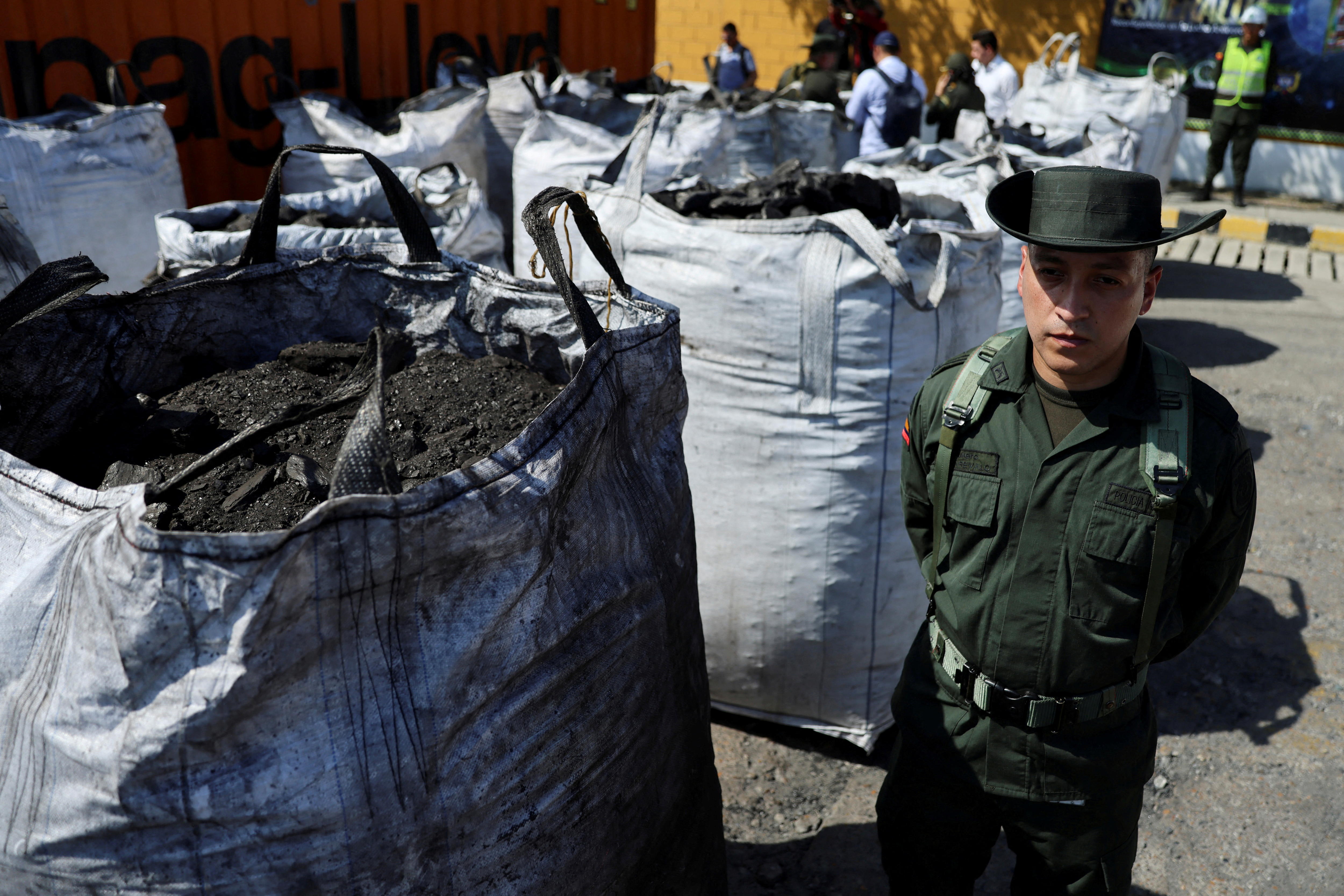 A Colombian policeman in dark green clothing and a hat standing next to large upright white bags filled with charcoal.