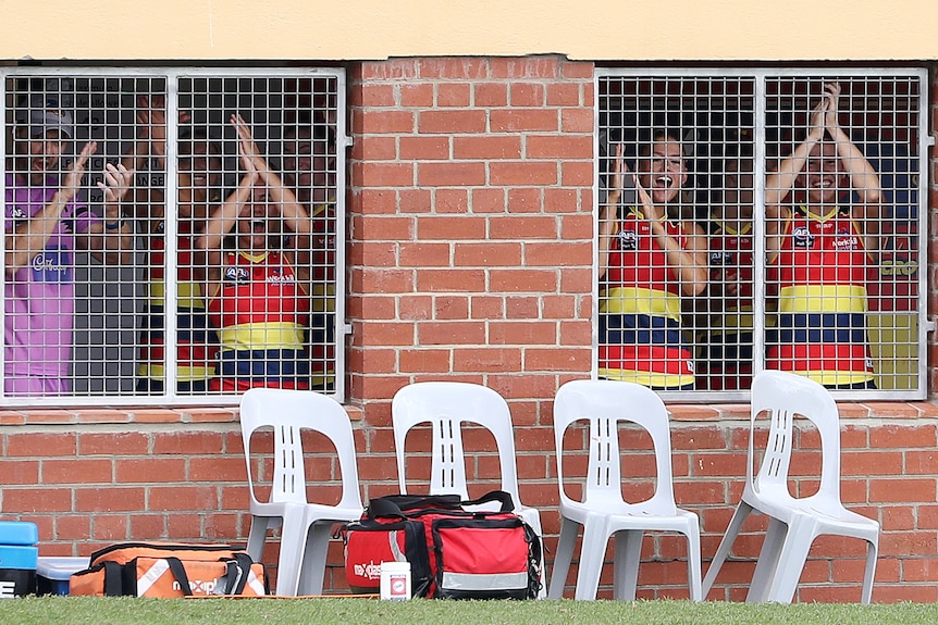 Adelaide Crows AFLW players applaud from behind metal bars on windows at Norwood Oval.